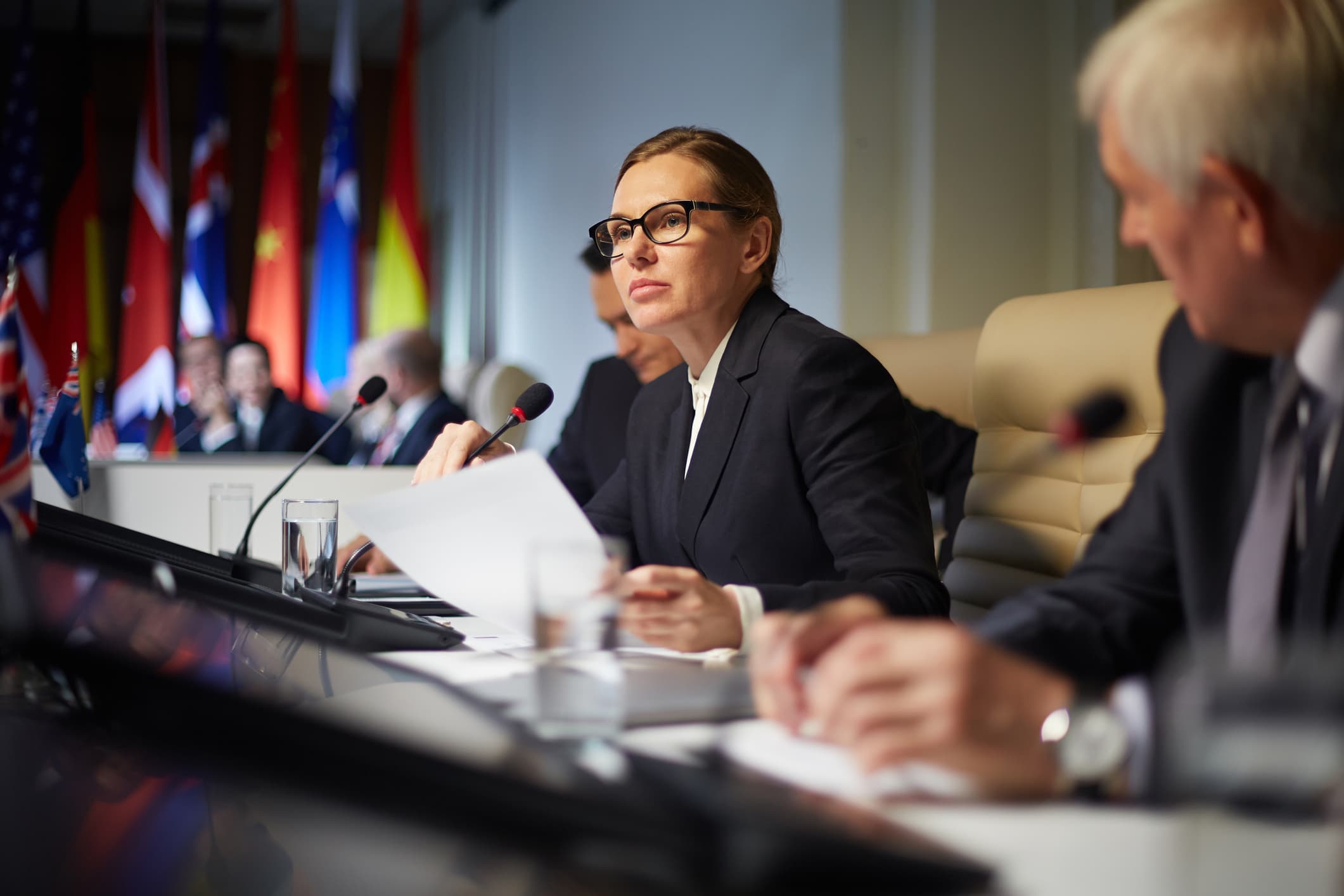 A woman in business attire wearing glasses at the table with political leaders and world flags behind them.