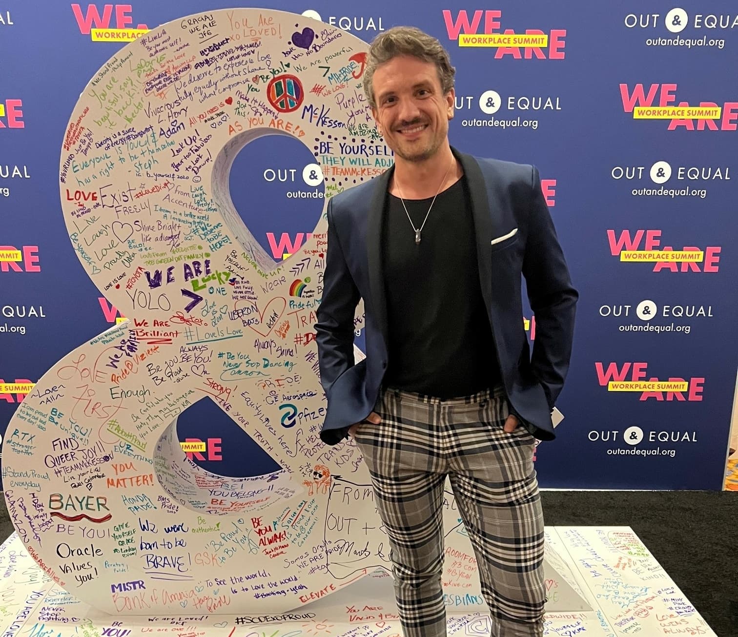 Weber Shandwick Q+ Chair-Elect John McCourt stands in front of a large white ampersand with various colored signatures at this year's Out & Equal conference.