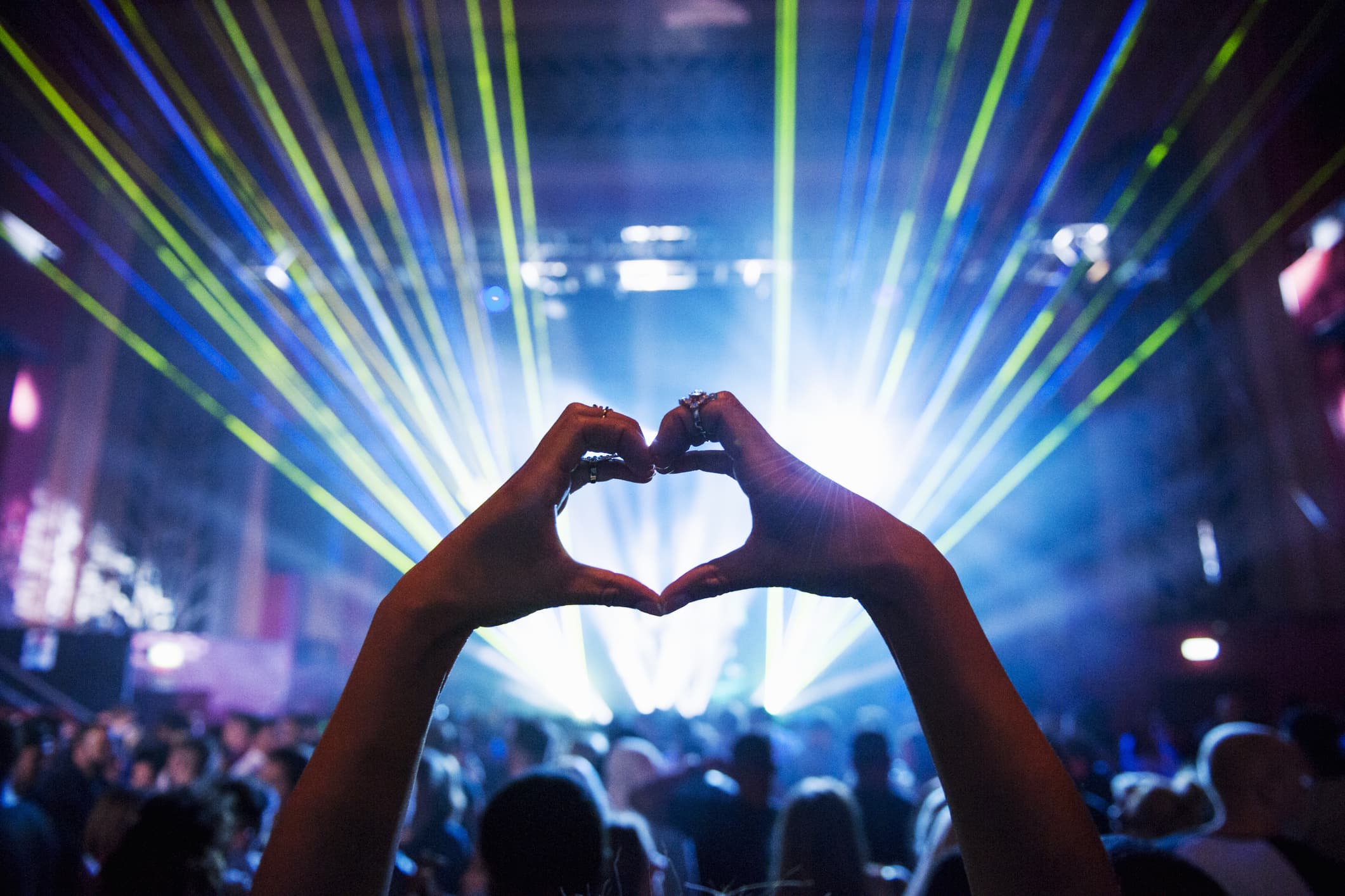 Woman making heart shape with hands at music event, as rays of light from the event lighting come out towards her in the audience