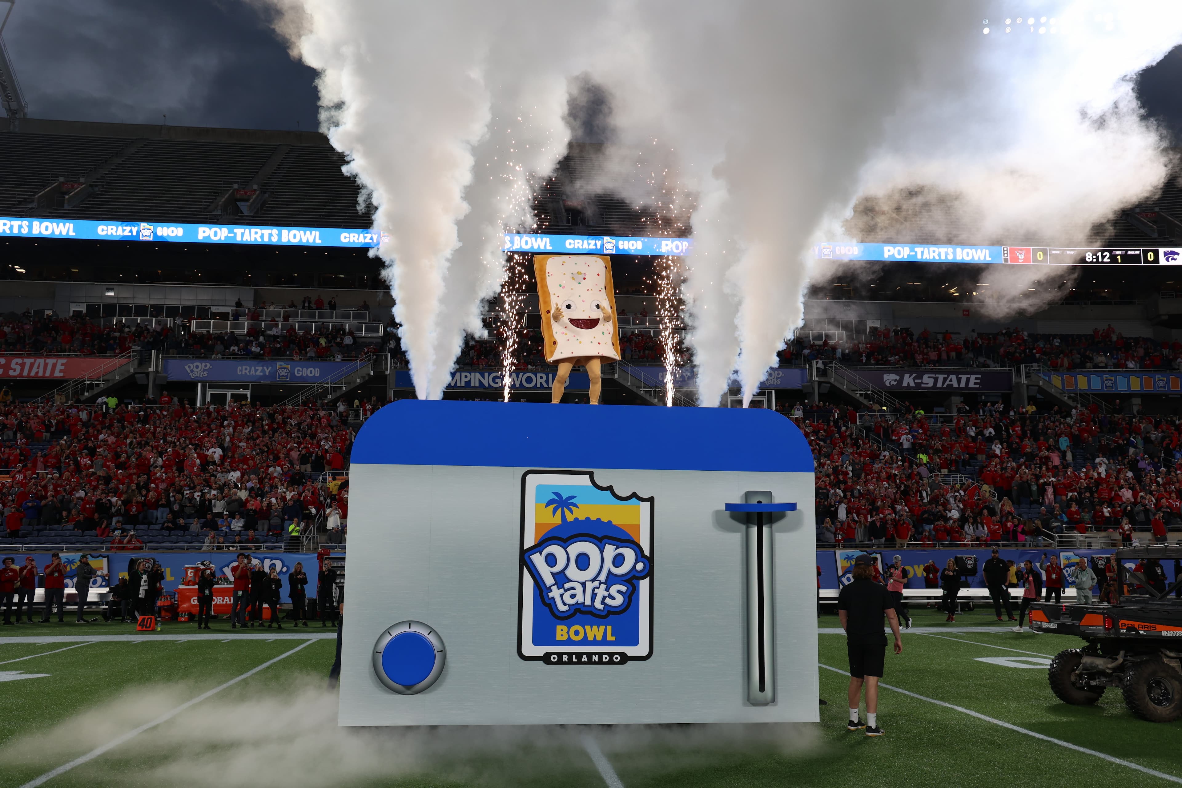 The Pop-Tarts mascot stands on top of a giant Pop-Tart branded toaster oven with smoke coming out to either side of them and a full stadium of people around them