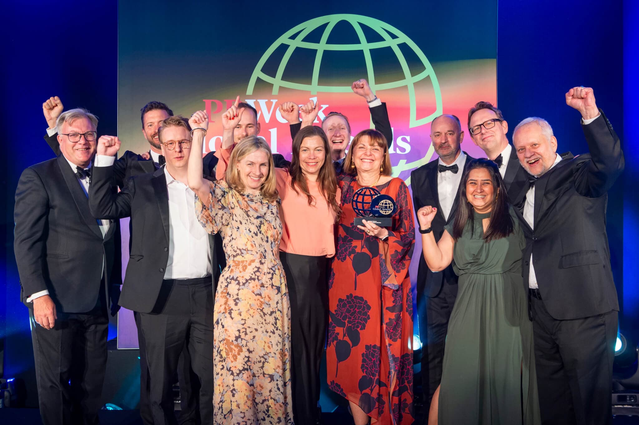 12 people stand on stage at the 2024 PRWeek Global Awards with their hands raised high, smiling as they cheer.