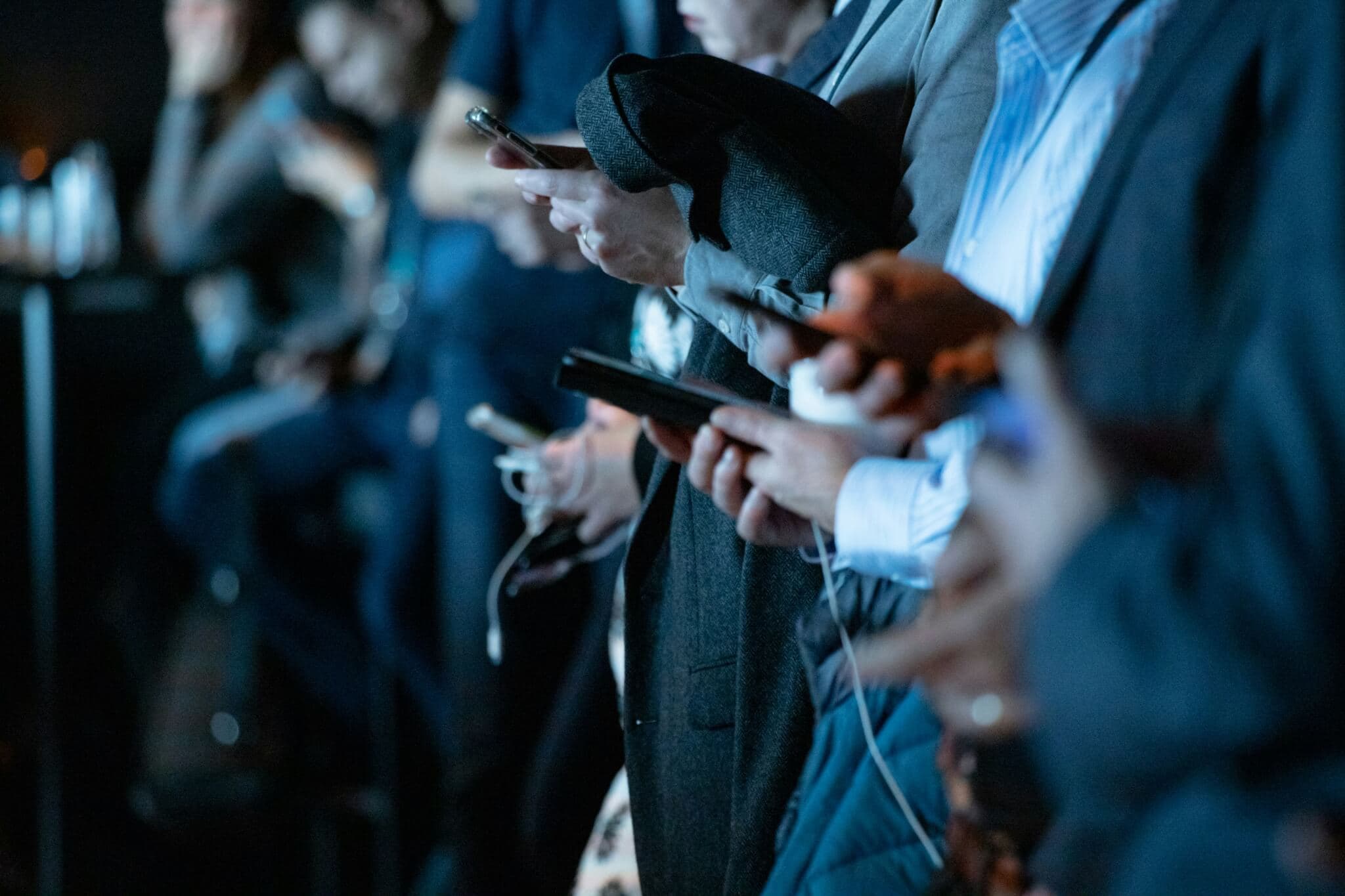 Multiple people stand in a row in a dark room, with their hands down scrolling on their smartphones