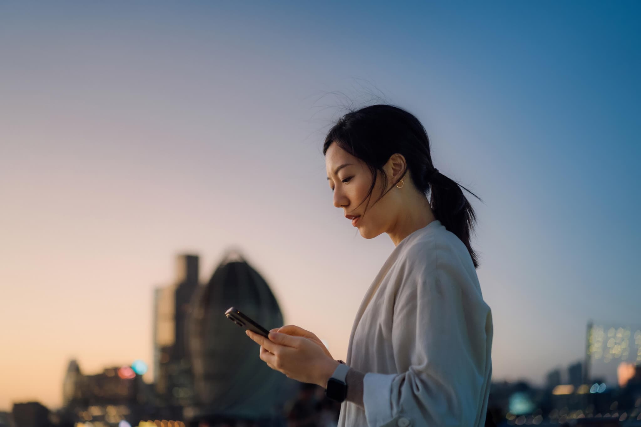 Modern young Asian woman walking in the city text messaging on smartphone on the go, against urban city scene with corporate skyscrapers at sunset. Lifestyle and technology