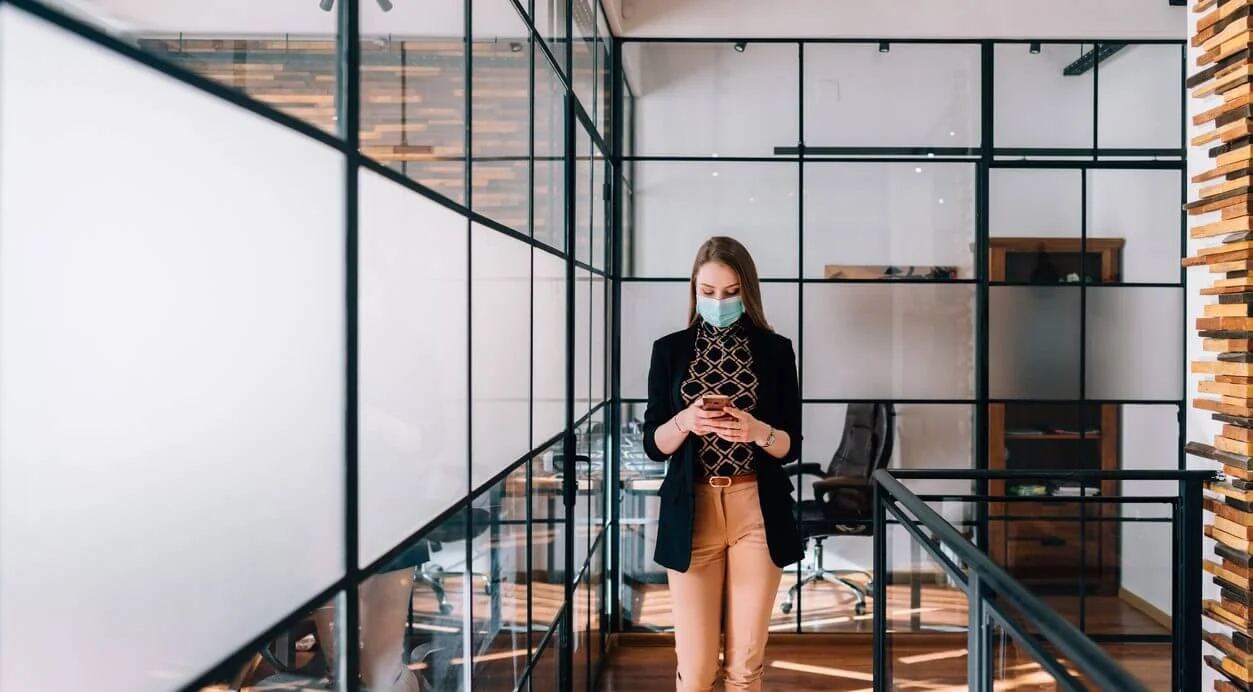 Professional woman wearing a face mask using a smartphone in a modern office corridor, reflecting workplace changes during COVID-19 return-to-work transition